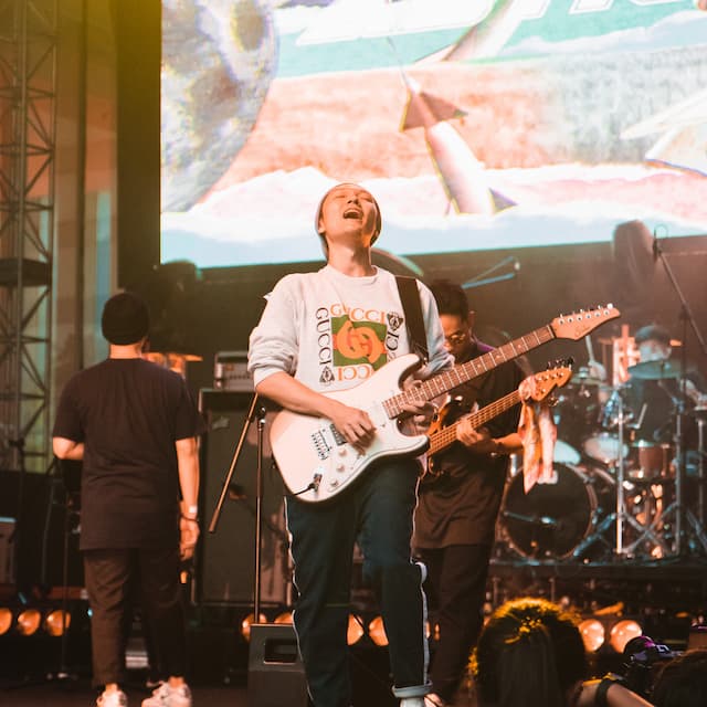 A man closes his eyes and smiles while playing guitar in front of a large screen